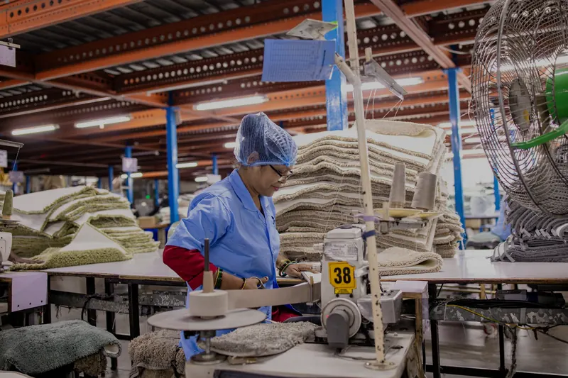 Bir tekstil fabrikasında bone takmış bir çalışan endüstriyel dikiş makinesinde ürün kenarı dikiyor; arkada istiflenmiş tekstil ürünleri
