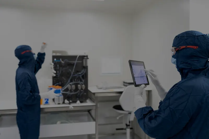 Mavi koruyucu tulum, başlık ve maske giymiş iki teknisyen bir laboratuvarda; biri cihazın başında çalışıyor, diğeri elindeki tablete bakıyor
