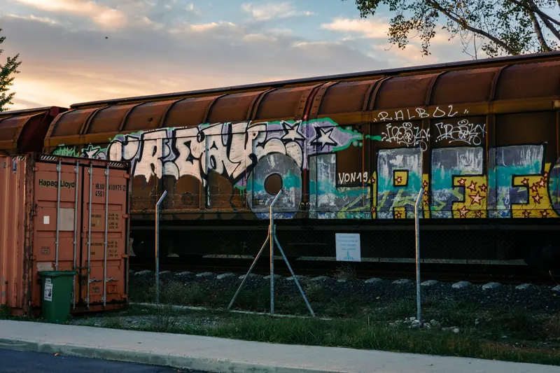 Çerkezköy'de gün batımında raylar üzerinde duran grafitili bir yük vagonu; solda bir konteyner, önde tel çit ve çakıllı demiryolu kenarı