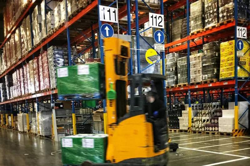 Yüksek raflarla dolu bir depoda koridorda ilerleyen sarı bir forklift ve paletli yükler