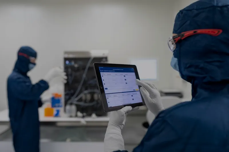Temiz oda kıyafetli bir teknisyen elindeki tablette elektronik parti kaydı yazılımını kullanıyor; arkada başka bir teknisyen çalışıyor