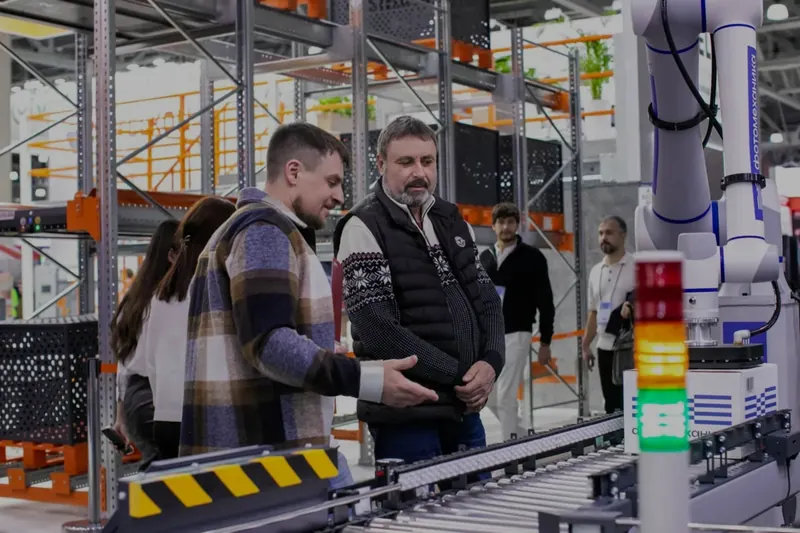 Bir fuarda depo otomasyonu için kurulmuş konveyör hattını ve robot kolu inceleyen ziyaretçiler