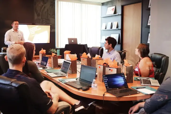Presenter at a screen addressing a team with laptops around a conference table – business development