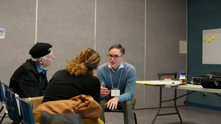 Trainer wearing a name badge explains a topic to two seated participants – training and certification